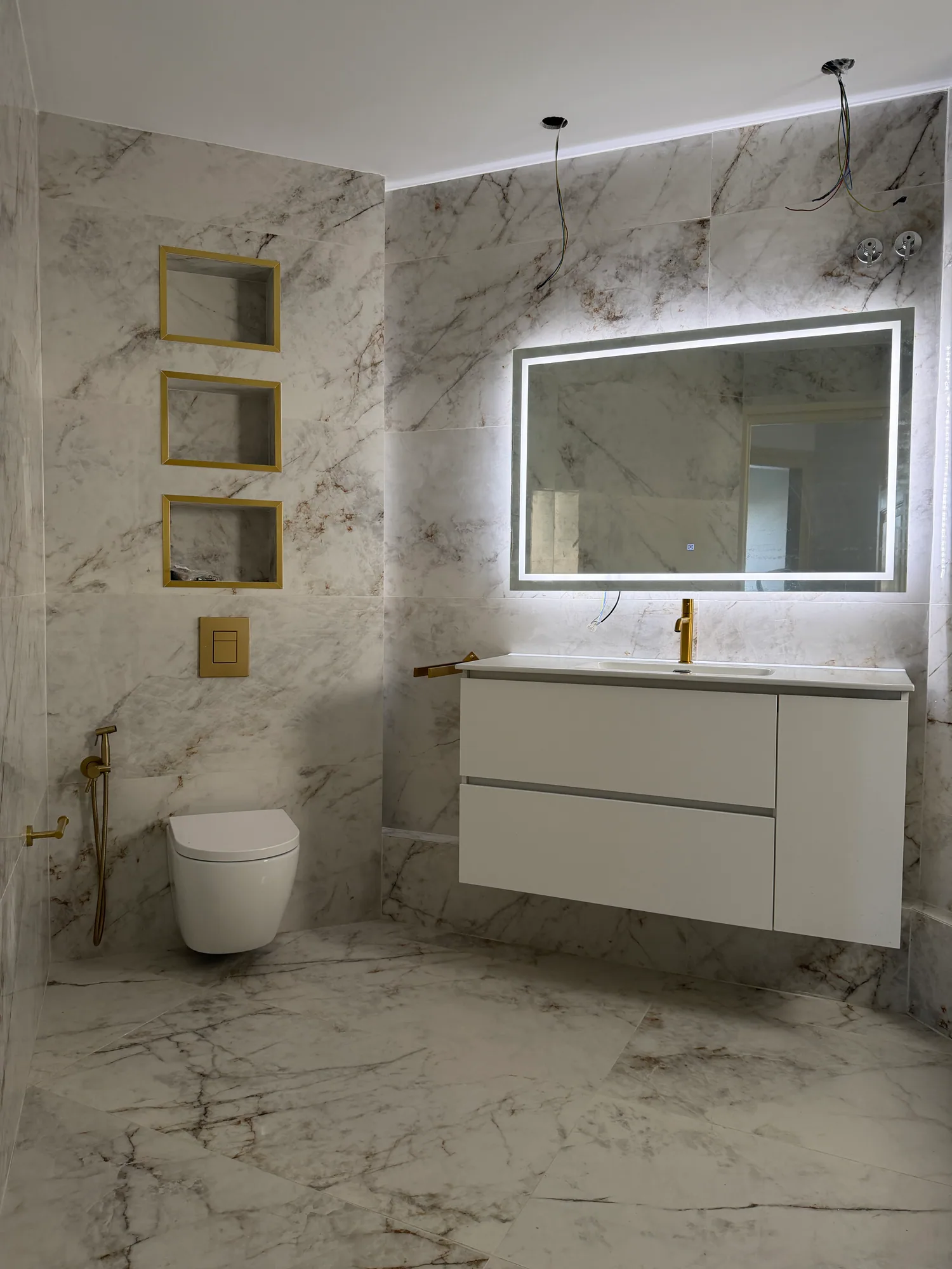 Baño revestido de mármol, con espejo retroiluminado, mueble suspendido y griferías doradas